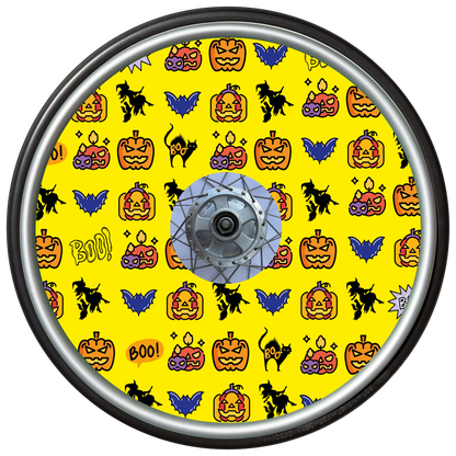 Wheelchair wheel cover on a wheel featuring pumpkins, bats, witches and Boo! on a yellow background.