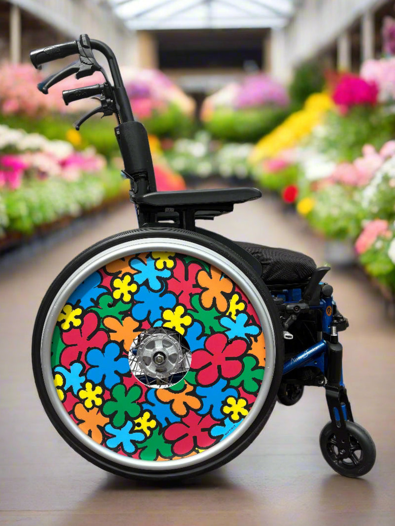 Wheelchair with a colorful, cartoon flowers wheel cover in red, orange, green, yellow and blue, set in a garden centre with blurred flower background. 