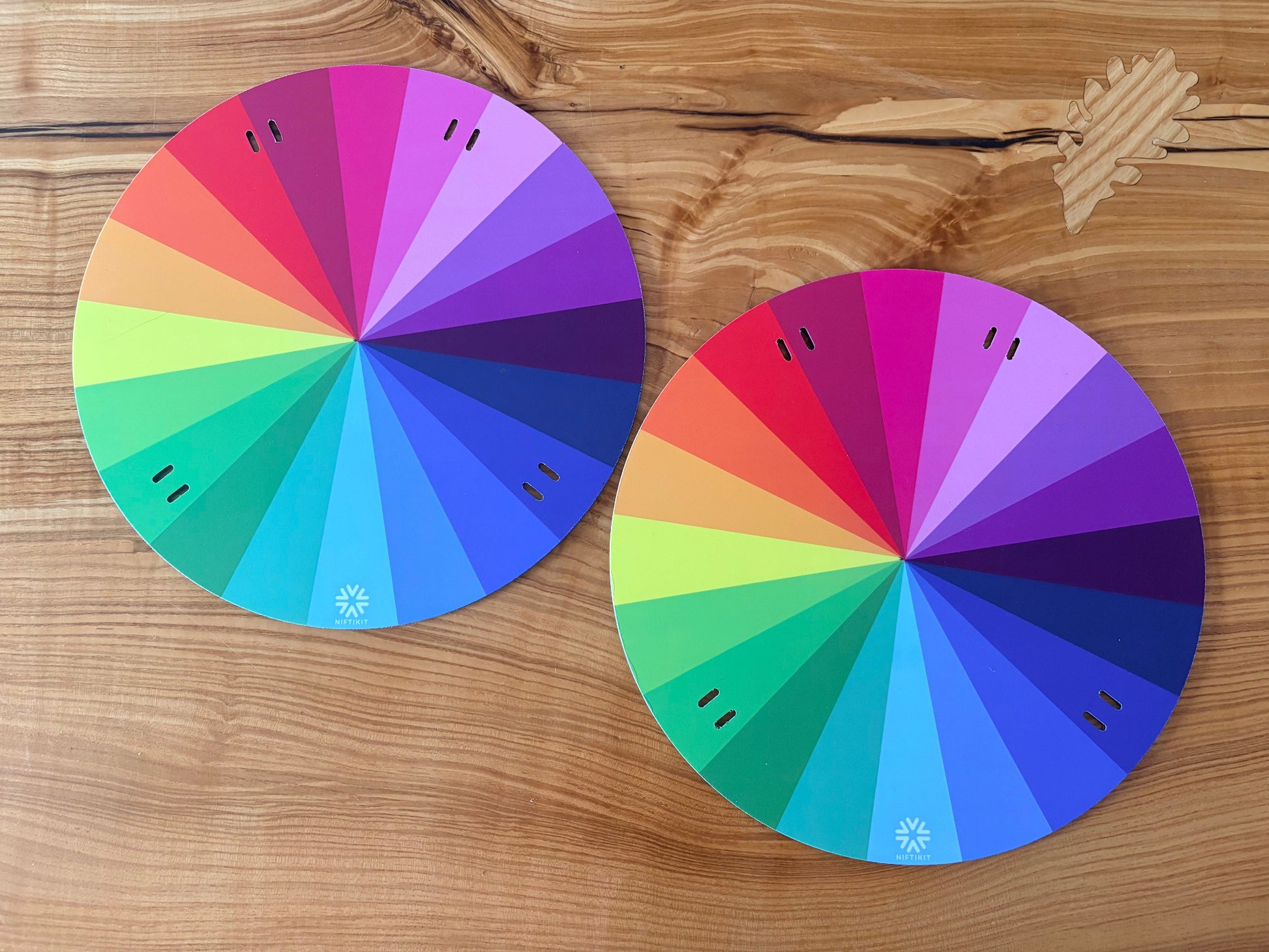 Pair of 320mm Niftikit Wheelchair Wheelcovers with rainbow gradient segments placed on a wooden surface.