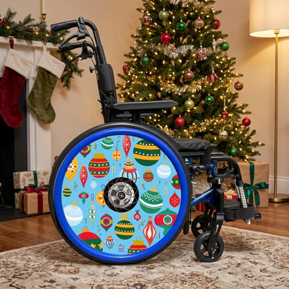 A black manual wheelchair with a light blue and multicolor Christmas ornament pattern spoke guard on the large rear wheel, positioned in a cozy living room setting with a decorated Christmas tree, fireplace, stockings, and wrapped gifts in the background.
