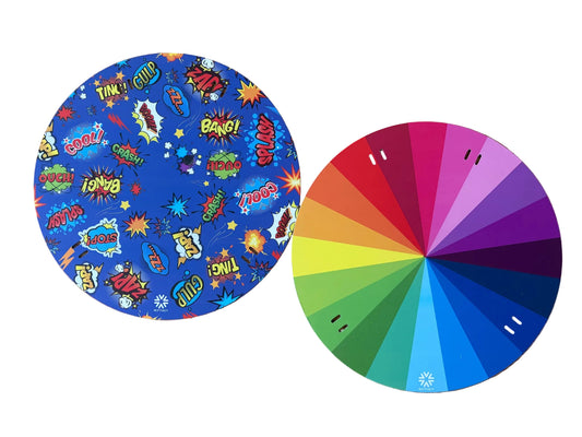 Reversible 320mm Niftikit Wheelchair Spoke Guards, features a colorful comic-style pattern with words like 'BOOM', 'ZAP!', and 'SPLASH' on a blue background, and the other shows a rainbow color wheel with gradient segments from green and yellow to red, orange, pink, purple, and blue.