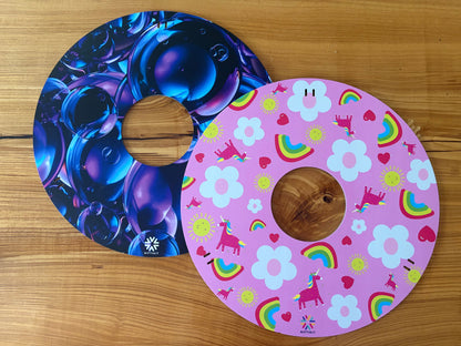 Pair of wheelchair wheelcovers, one with blue bubble design and the other with pink unicorn, floral and rainbow design, on a wooden surface.