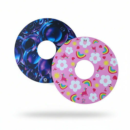 Pair of wheelchair wheelcovers, one with blue bubble design and the other with pink unicorn, floral and rainbow design, on a wooden surface.