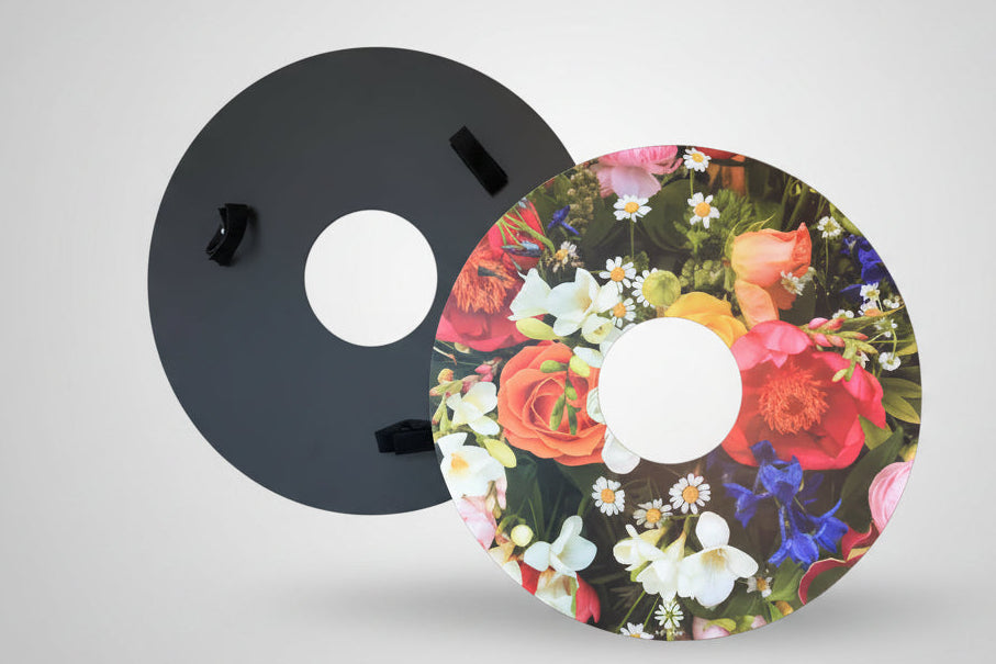 One pair of NIftikit wheelchair wheel covers. One showing the back of the spoke guard, with three riveted Velcro straps on a black disc. The other showing the front of the spoke guard showing a bright floral design. 